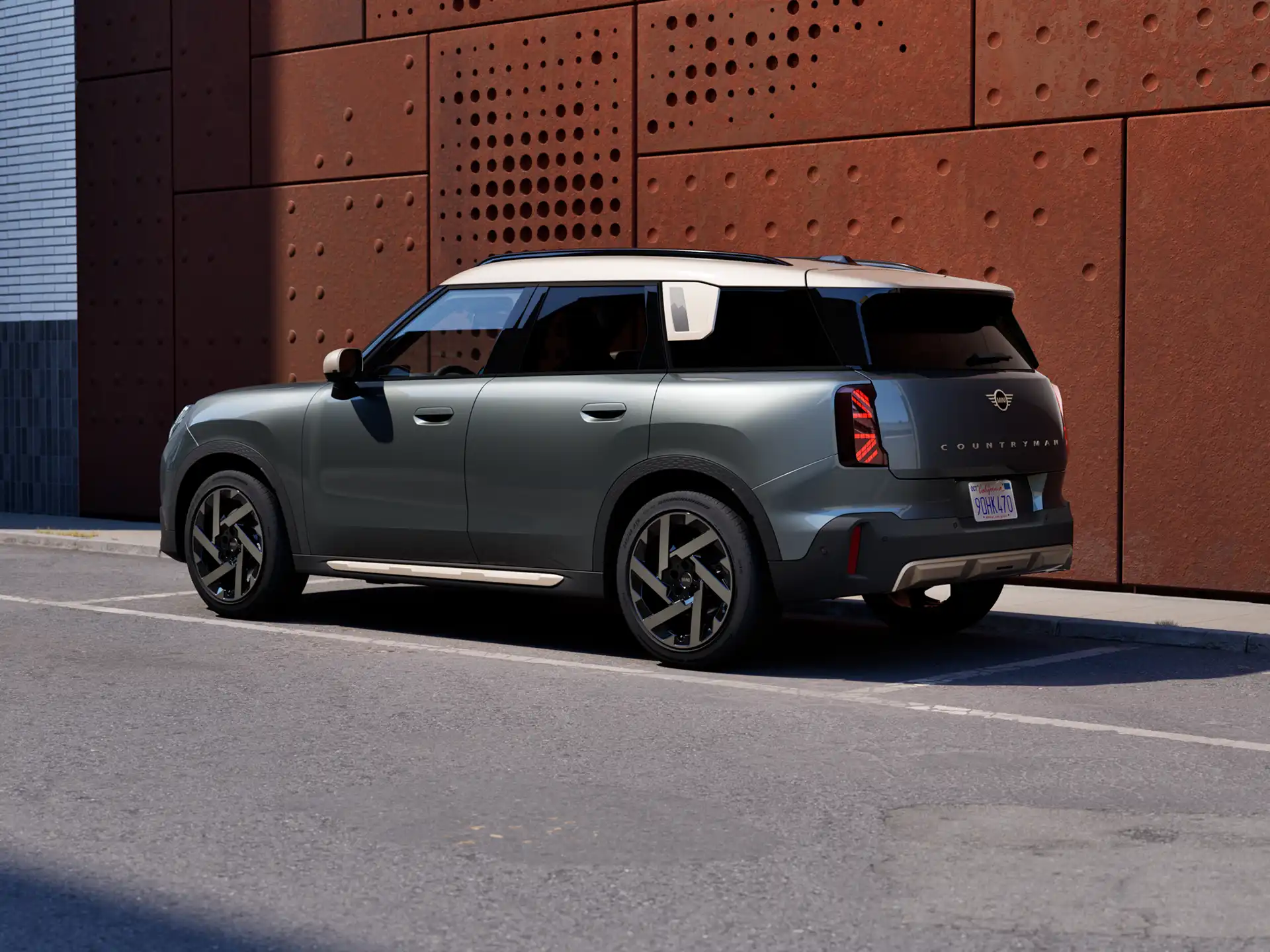 Rear three‑quarter view of a MINI Countryman in Sage Green Metallic parked beside a rust‑coloured wall.
