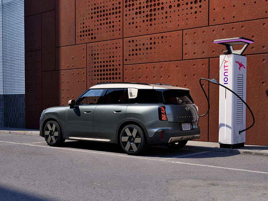 Side view of a MINI Countryman parked on a city street while charging at an IONITY electric charging station beside a perforated rust‑coloured wall.