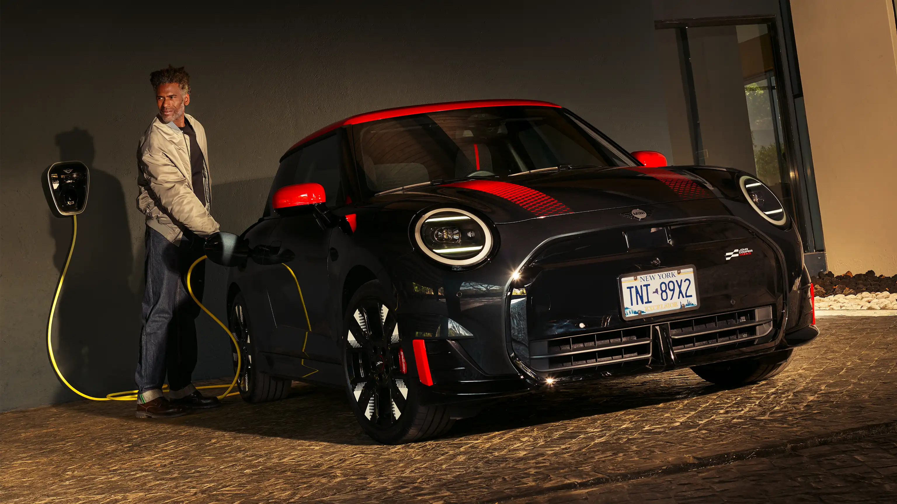 Person plugging in a MINI John Cooper Works in Midnight Black II with Chili Red roof at a home charging station